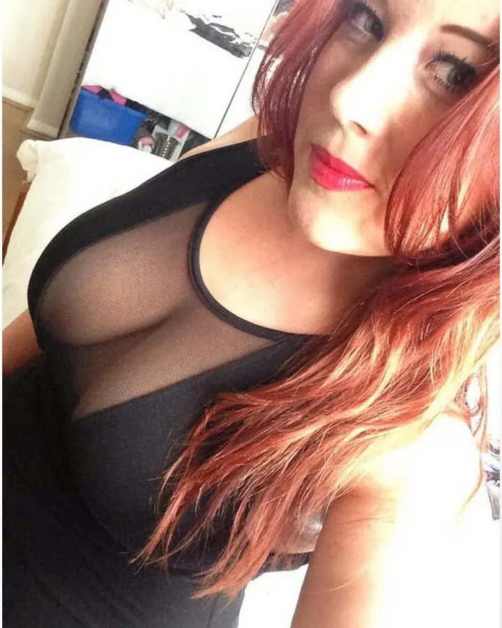 Femme aux cheveux roux, en lingerie noire, regardant vers le bas, dans une pièce sombre.
