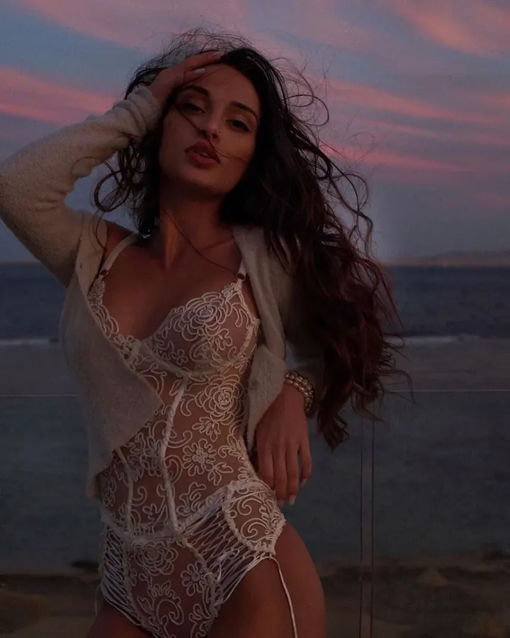 Femme vue de dos, en robe noire transparente scintillante, portant des talons hauts, dans une chambre.
