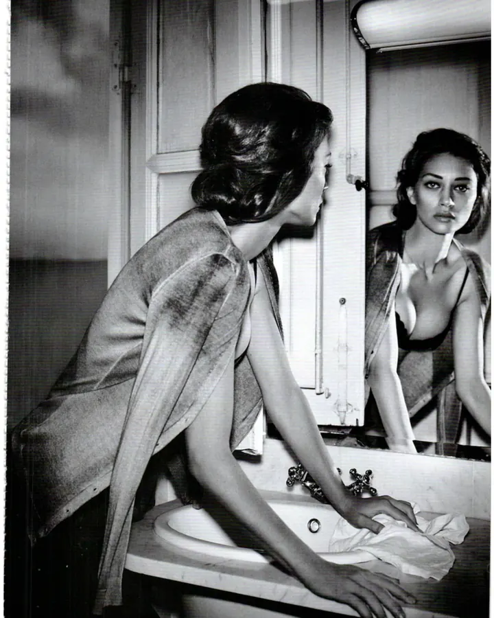 Femme en noir et blanc, penchée sur un lavabo, regardant son reflet dans le miroir, vêtue d’un peignoir et d’un soutien-gorge.