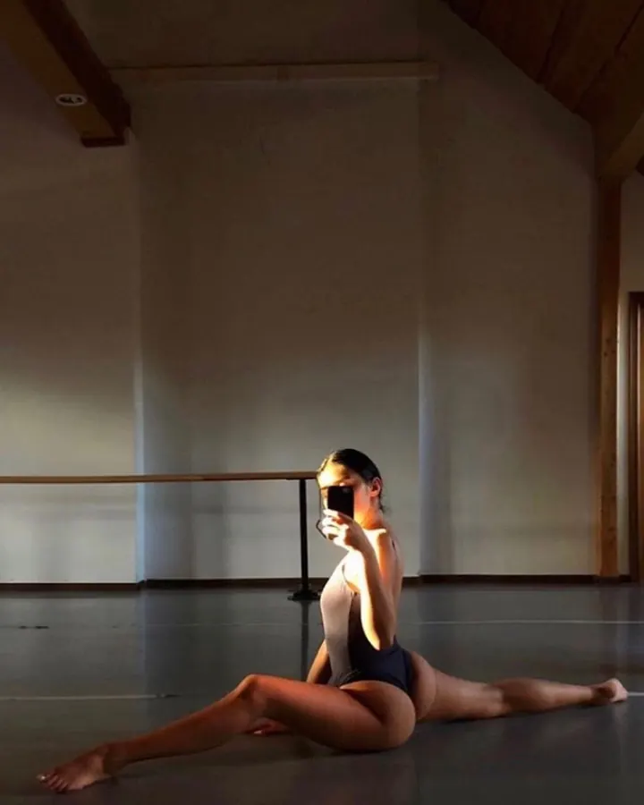 Femme en justaucorps noir et blanc, en train de faire la splits dans une salle de danse avec barre et miroir.