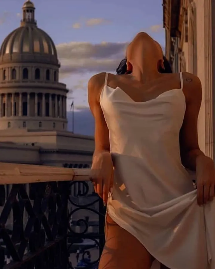 Femme en haut blanc, regardant vers le ciel, debout sur un balcon avec une vue sur un bâtiment à dôme au coucher du soleil.
