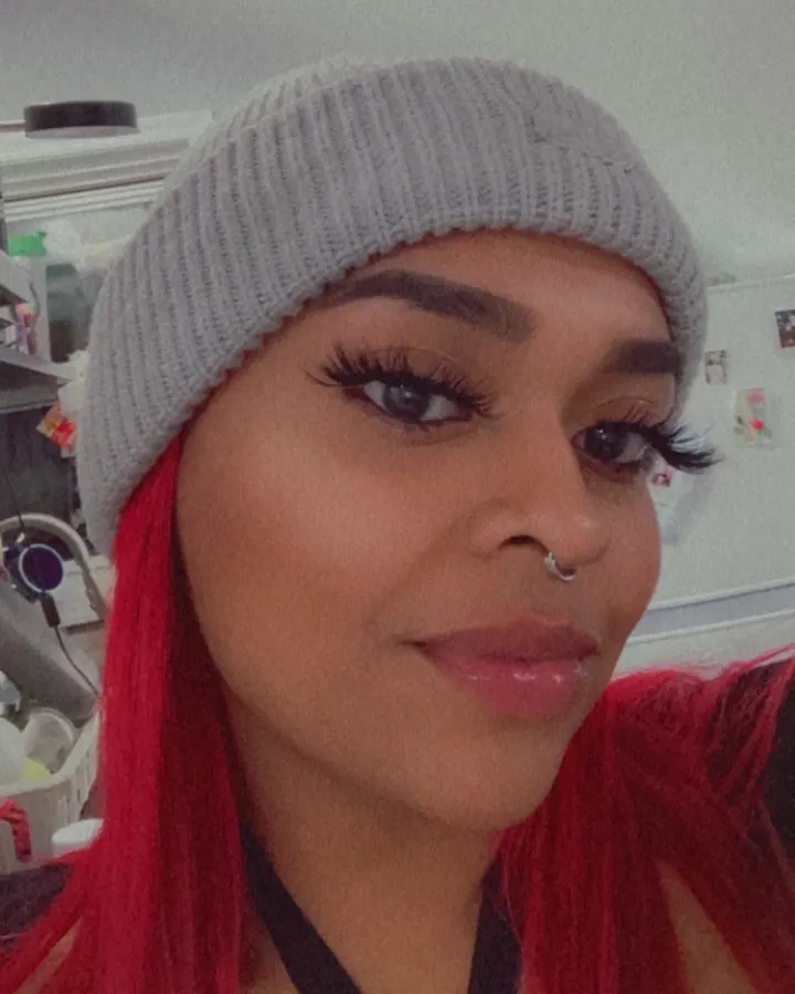Femme aux cheveux roux en lingerie noire, posant sur un lit rouge.