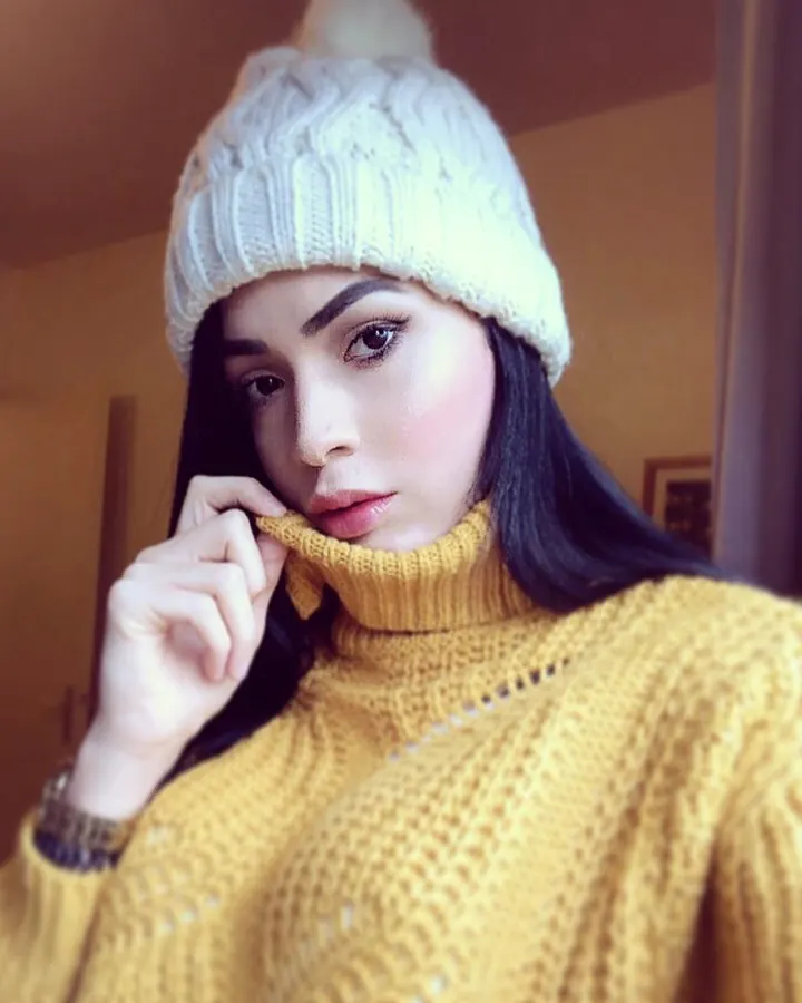 Femme aux cheveux noirs, bonnet blanc à pompon, pull jaune tricoté, main sur le col, regard intense