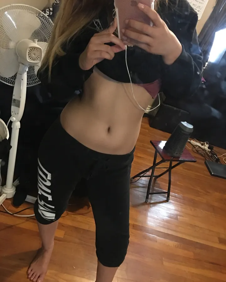 Femme en sweat noir, ventre découvert, tenant un téléphone rose, pieds nus sur parquet
