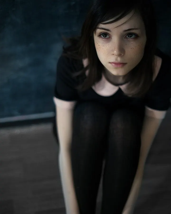 Femme aux cheveux bruns foncés, vêtue d’un haut noir, assise sur un sol en bois, les genoux repliés, regardant directement l'appareil avec un regard intense.