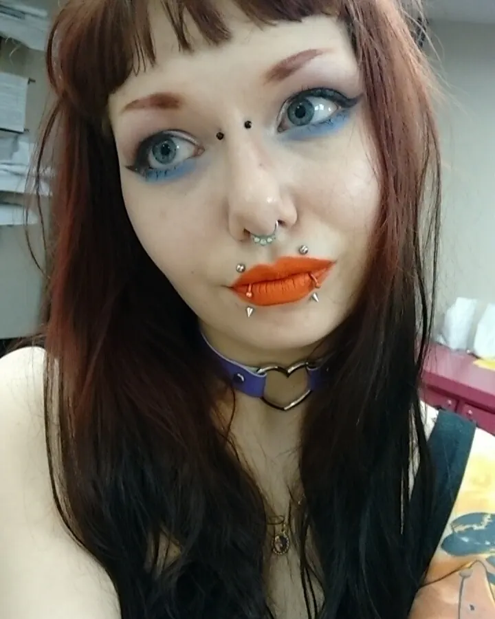 Femme aux cheveux bruns foncés avec frange, maquillage prononcé aux yeux bleus, lèvres orange vif, piercings multiples au visage, collier en cuir violet avec cœur métallique.