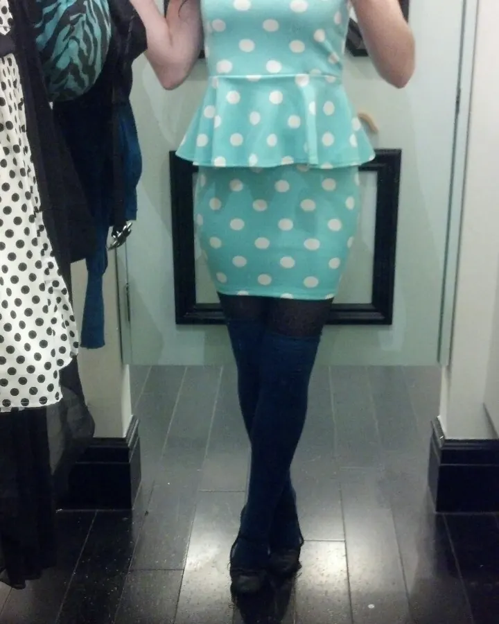 Femme européenne aux cheveux bruns-noirs, portant une robe turquoise à pois blancs, des collants noirs et des chaussures noires, prenant un selfie devant un miroir.