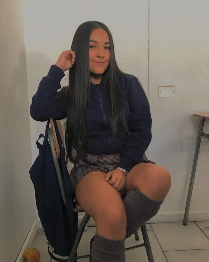 Jeune femme arabe aux cheveux bruns-noirs, portant un sweat bleu marine, une jupe à carreaux, des collants gris et des chaussettes hautes, assise sur une chaise.