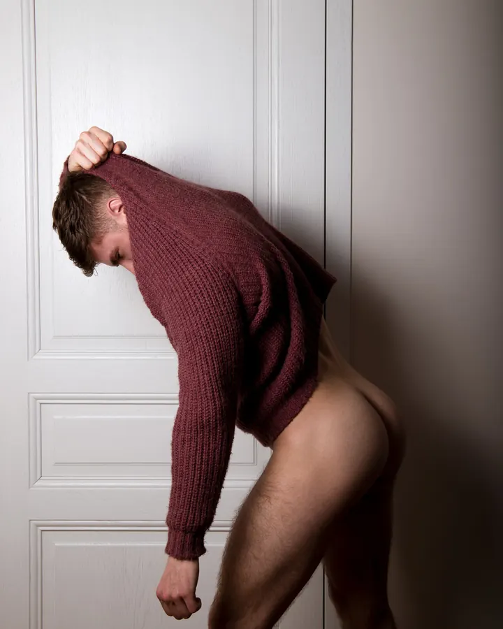 Homme en pull rouge se tenant penché contre une porte blanche, nu de la taille vers le bas.