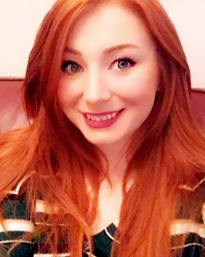 Femme aux cheveux roux portant un pull rayé vert et blanc, souriant, assise sur un canapé rouge.