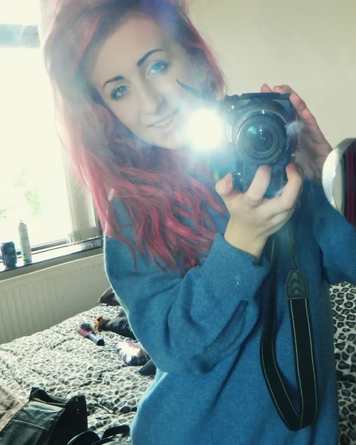 Femme aux cheveux roses tenant un appareil photo, souriant, en pull bleu, dans une chambre avec un lit à motif léopard.