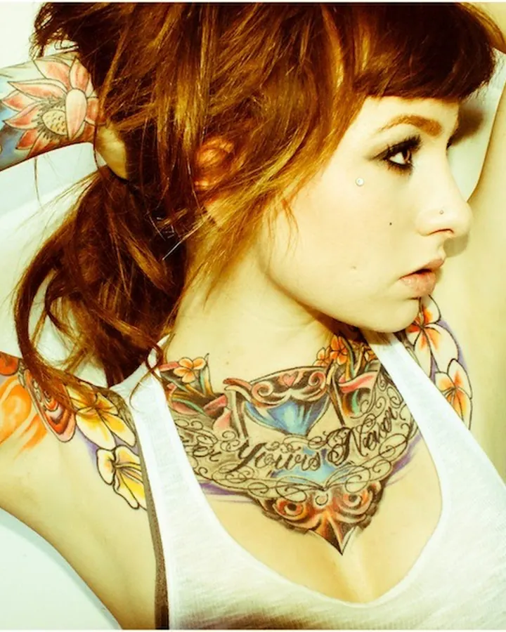 Femme européenne aux cheveux roux, tatouages colorés sur le bras et le cou, en débardeur blanc, regardant de côté, main dans les cheveux.