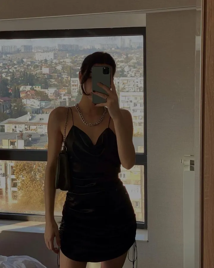 Femme européenne en robe noire en cuir, prenant un selfie devant une fenêtre avec vue sur une ville, sac à bandoulière, collier en chaîne.