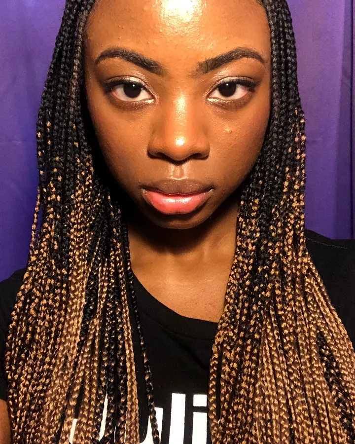 Femme africaine aux cheveux longs noirs, en t-shirt noir, regardant la caméra de face, visage net, cheveux tressés avec mèches blondes.