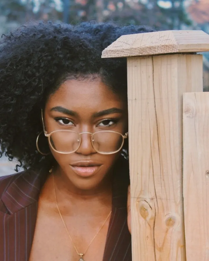 Femme africaine aux cheveux bouclés noirs, lunettes rondes dorées, regardant la caméra depuis derrière une clôture en bois.