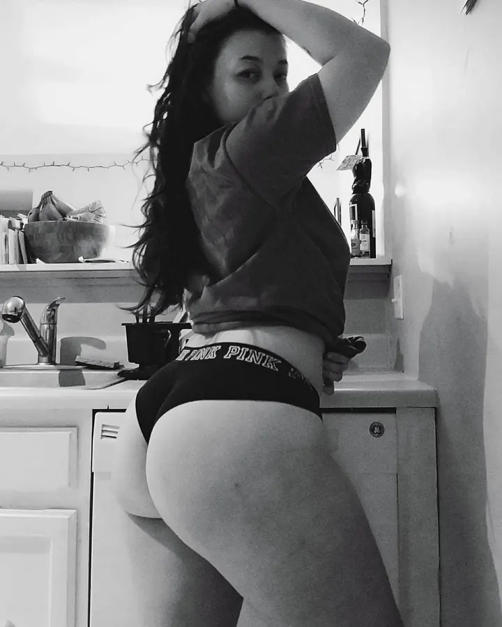 Photo en noir et blanc d'une femme portant un t-shirt et un string, les mains sur la tête, dans une cuisine.