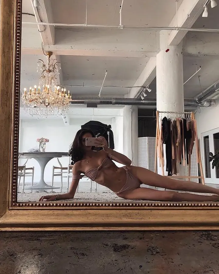 Femme aux cheveux bruns-noirs en lingerie blanche, posant de profil contre un mur bleu.