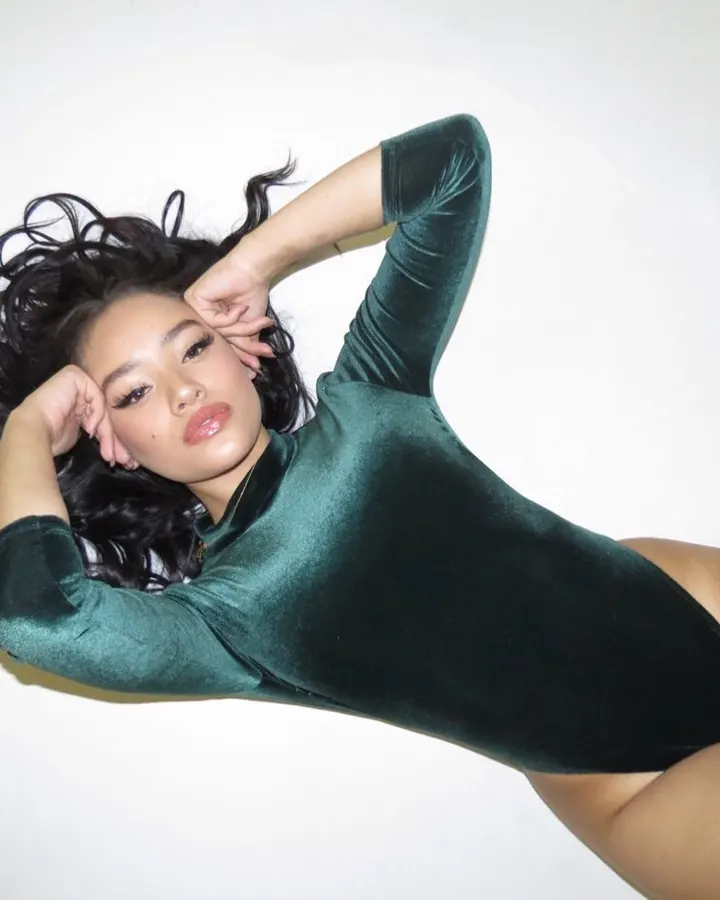 Femme aux cheveux noirs, portant un body vert velours, allongée sur le côté, les mains derrière la tête, sur fond blanc.