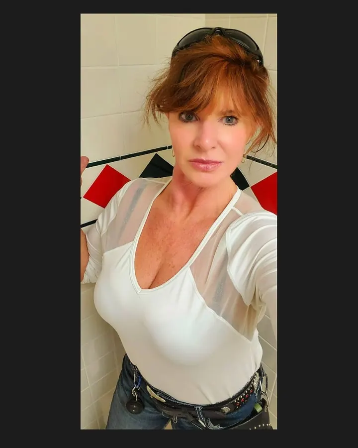 Femme aux cheveux roux, portant un haut blanc transparent, un jean, et un ceinturon décoré, prenant un selfie dans une salle de bain.