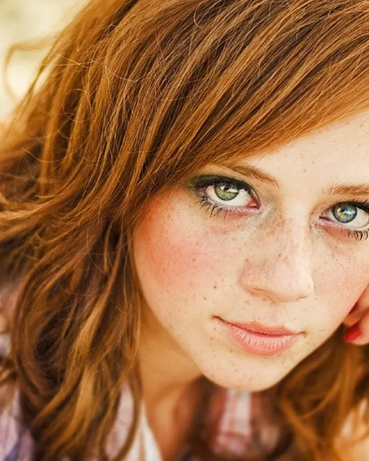 Femme aux cheveux roux et yeux verts, regardant l'objectif, la main posée sur la joue, vêtue d'une chemise à carreaux colorés.