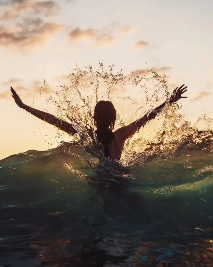 Silhouette féminine dans l'eau, bras ouverts, éclaboussures au coucher du soleil