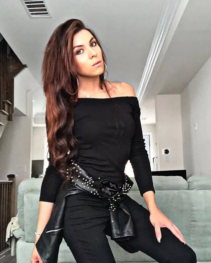 Femme en tenue noire, bustier, pantalon, ceinture en cuir noir ornée de clous, posant dans un salon moderne