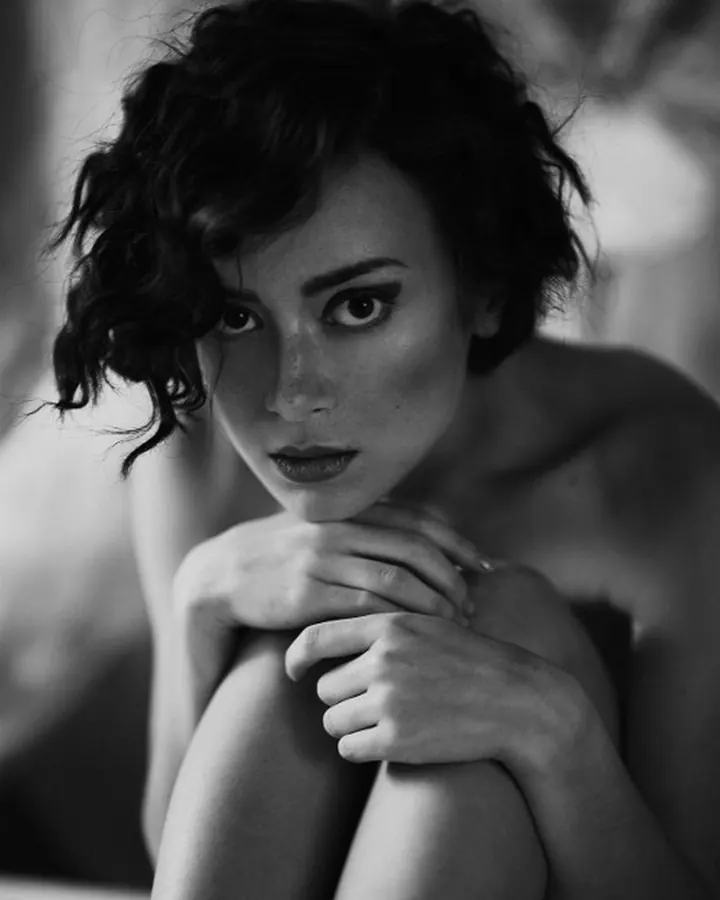 Femme en noir et blanc, cheveux courts bouclés, nue, les mains sur les genoux, regardant l'appareil