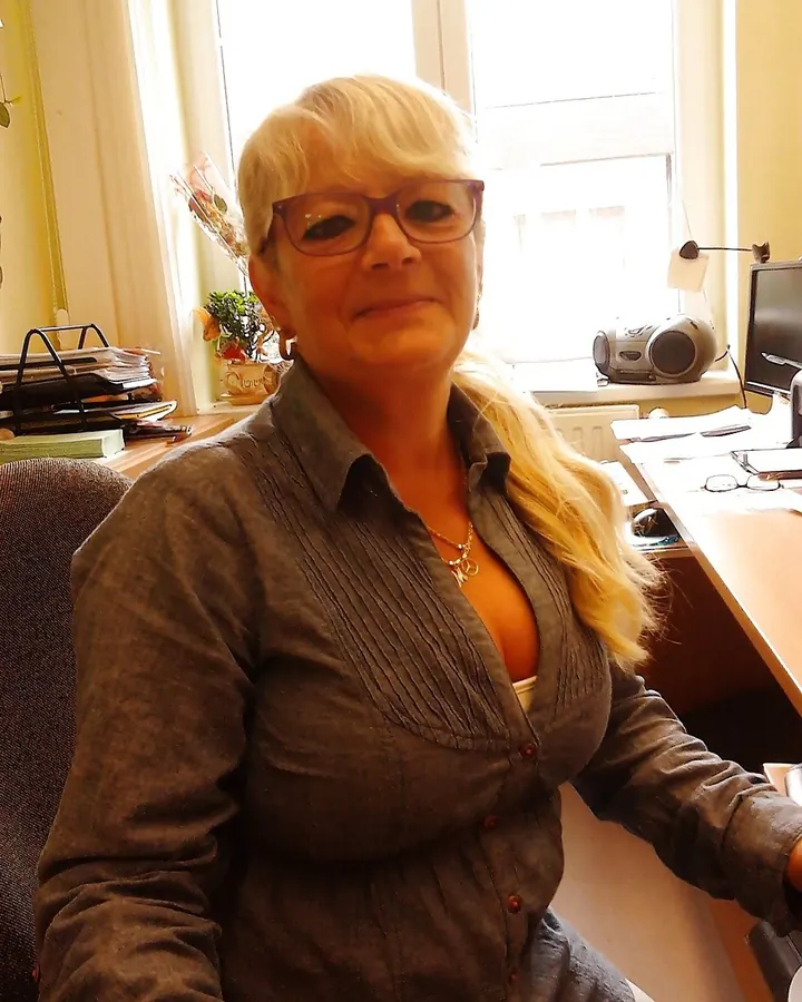 Femme blonde assise à un bureau, lunettes, chemise grise, regardant l'appareil