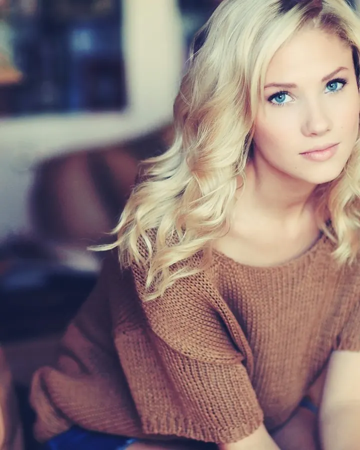 Femme blonde en pull marron, assise sur un canapé, regardant l'appareil, ambiance chaleureuse