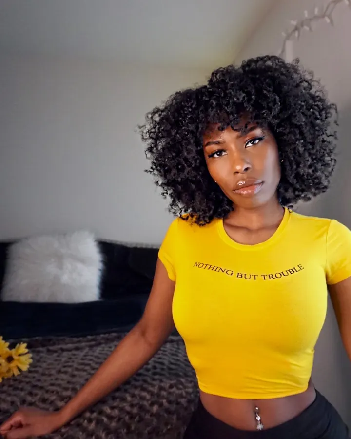 Femme africaine aux cheveux bouclés noirs, vêtue d'un t-shirt jaune avec l'inscription 'NOTHING BUT TROUBLE', posant devant un lit avec des fleurs jaunes.