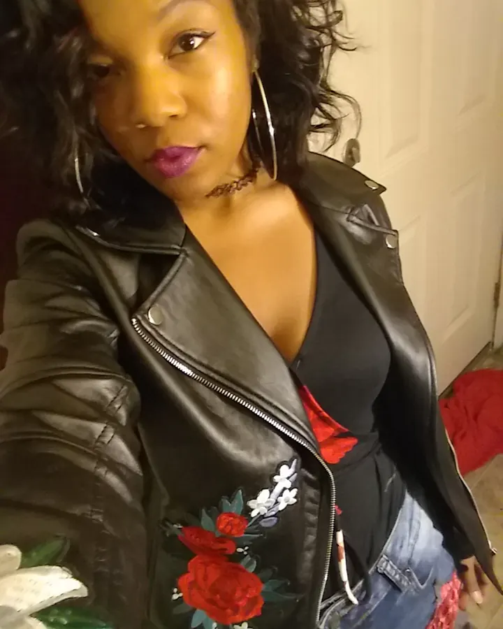 Femme africaine aux cheveux noirs ondulés, portant une veste en cuir noir avec des broderies de roses rouges, un collier fin, et des boucles d'oreilles grandes, regardant directement l'appareil.