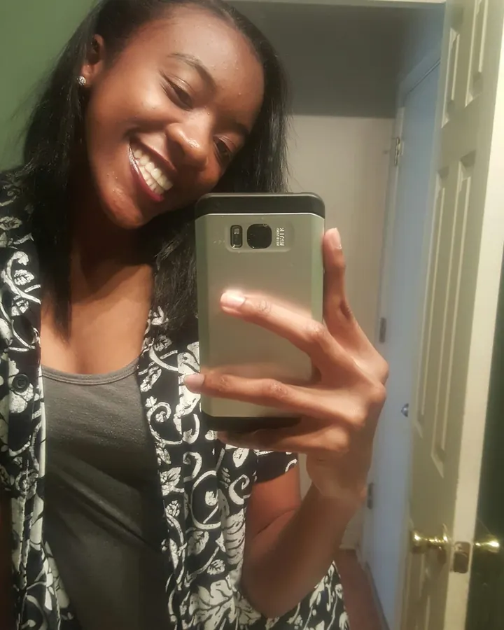 Femme africaine souriante prenant un selfie devant un miroir, vêtue d'un haut gris et d'une chemise à motifs