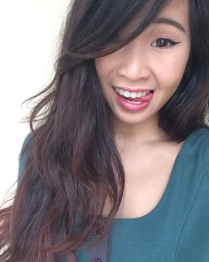 Femme asiatique souriante, cheveux longs ondulés, maquillage léger, pull vert foncé, fond blanc.