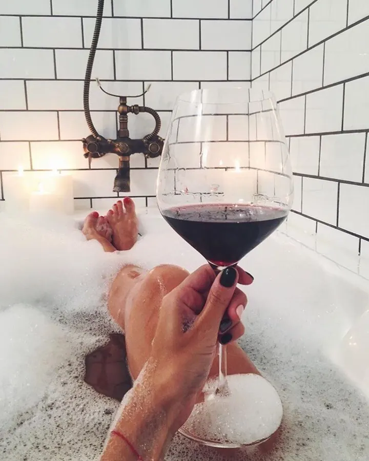 Pieds dans un bain moussant, main tenant un verre de vin rouge, ambiance éclairée par des bougies et carreaux blancs.