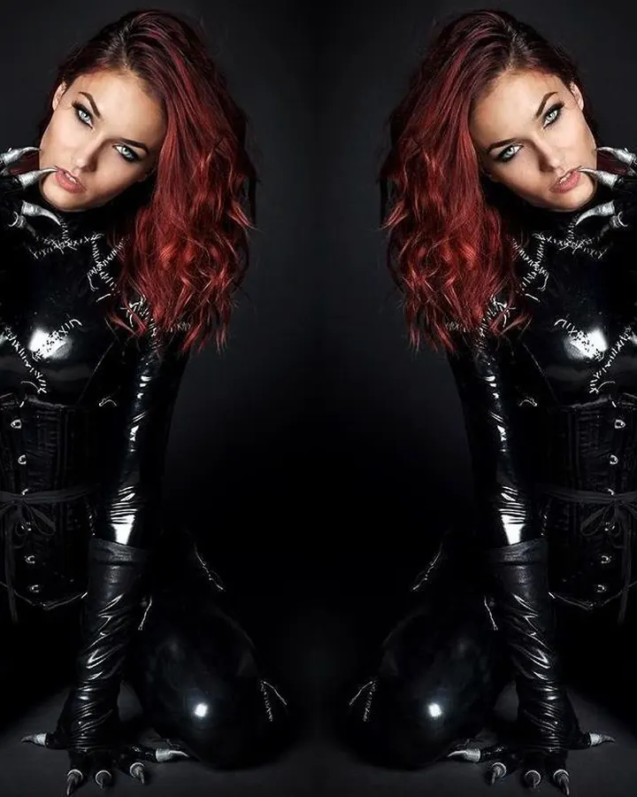 Femme aux cheveux roux, vêtue d'un costume noir brillant, agenouillée, regardant vers l'avant avec des gants noirs.
