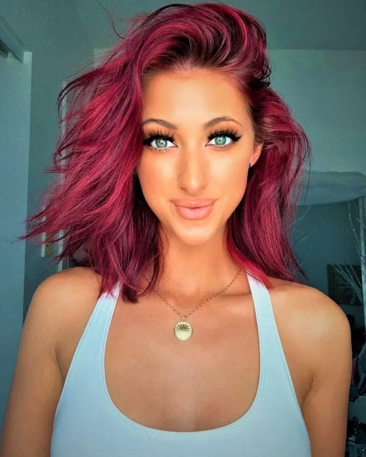 Femme aux cheveux roses et yeux verts en haut blanc, regardant l'appareil photo avec un sourire discret.