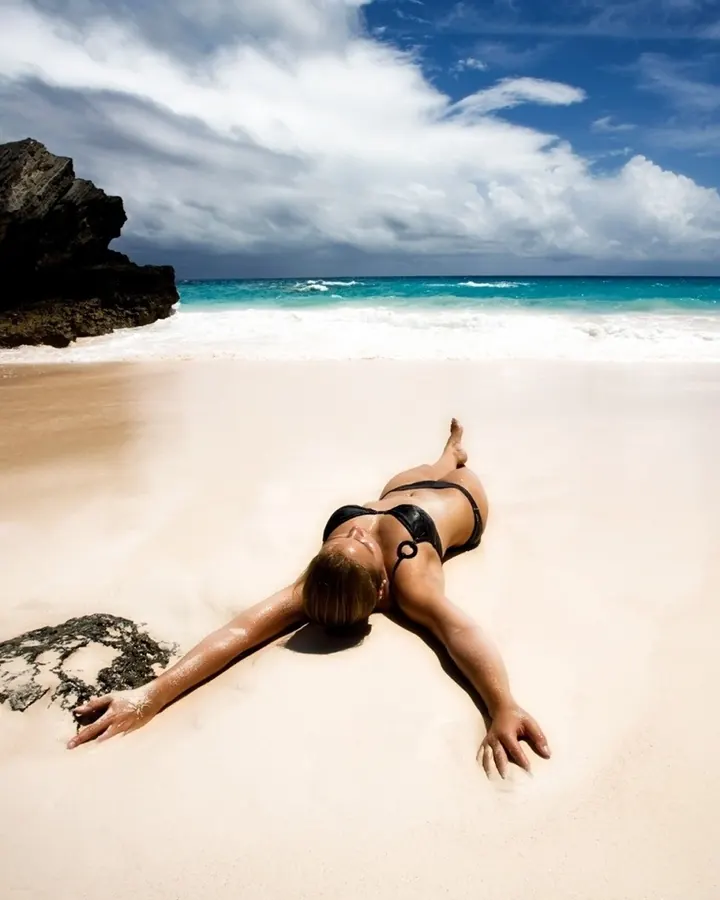Femme en maillot noir allongée sur le sable d'une plage, bras étendus, regardant vers le ciel.