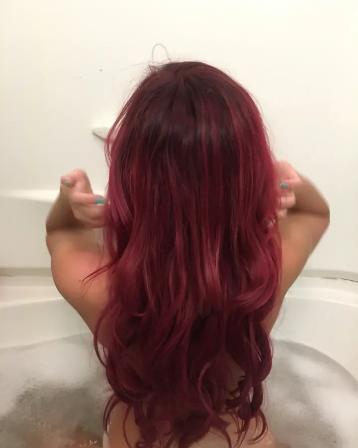 Femme aux cheveux rouges dans un bain moussant, vue de dos, les mains dans ses cheveux.