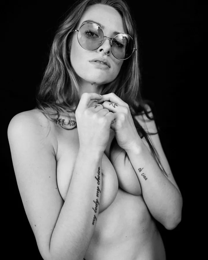Femme nue, sauf pour un string blanc et des bas blancs, assise sur un tapis, les mains sur ses seins, tatouages visibles.