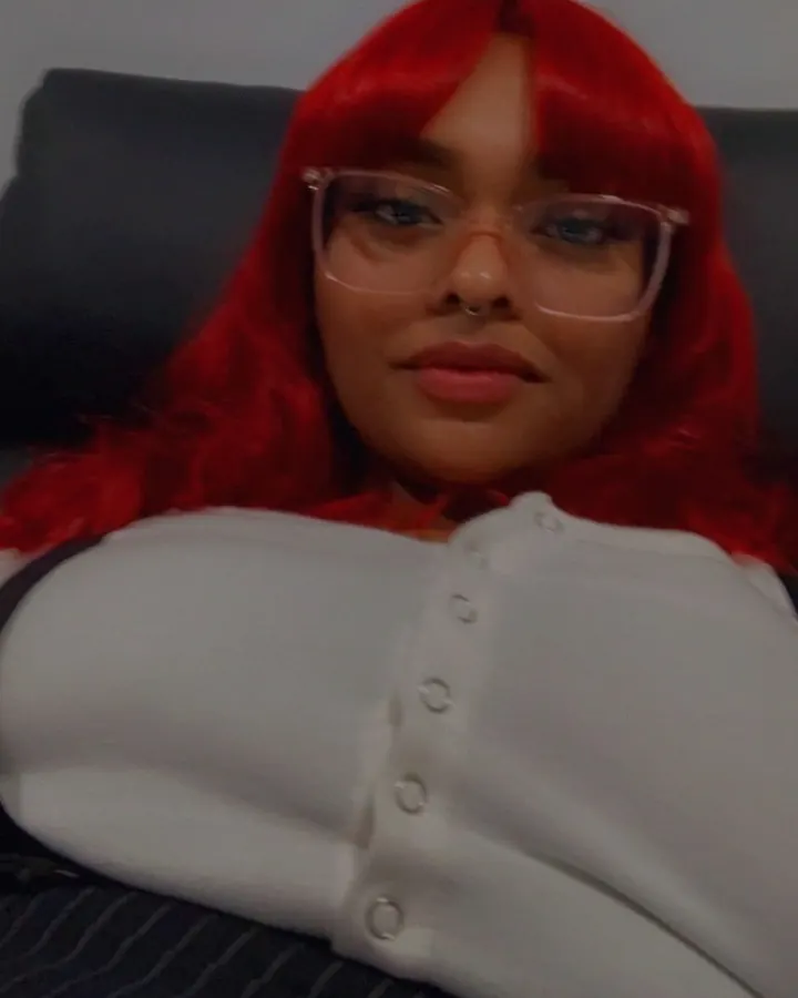Femme aux cheveux rouges en chemise blanche boutonnée, allongée sur un canapé, regardant l'appareil photo.