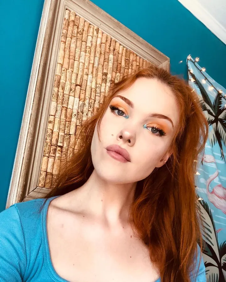Femme aux cheveux roux en lingerie rouge, avec des ailes blanches, se tenant devant un miroir.