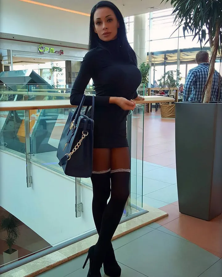Femme aux cheveux noirs en robe noire ajustée, tenant un sac à main, appuyée sur une rambarde dans un centre commercial.
