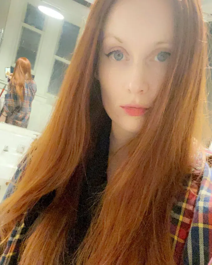Femme aux cheveux roux en chemise à carreaux, prenant un selfie devant un miroir.