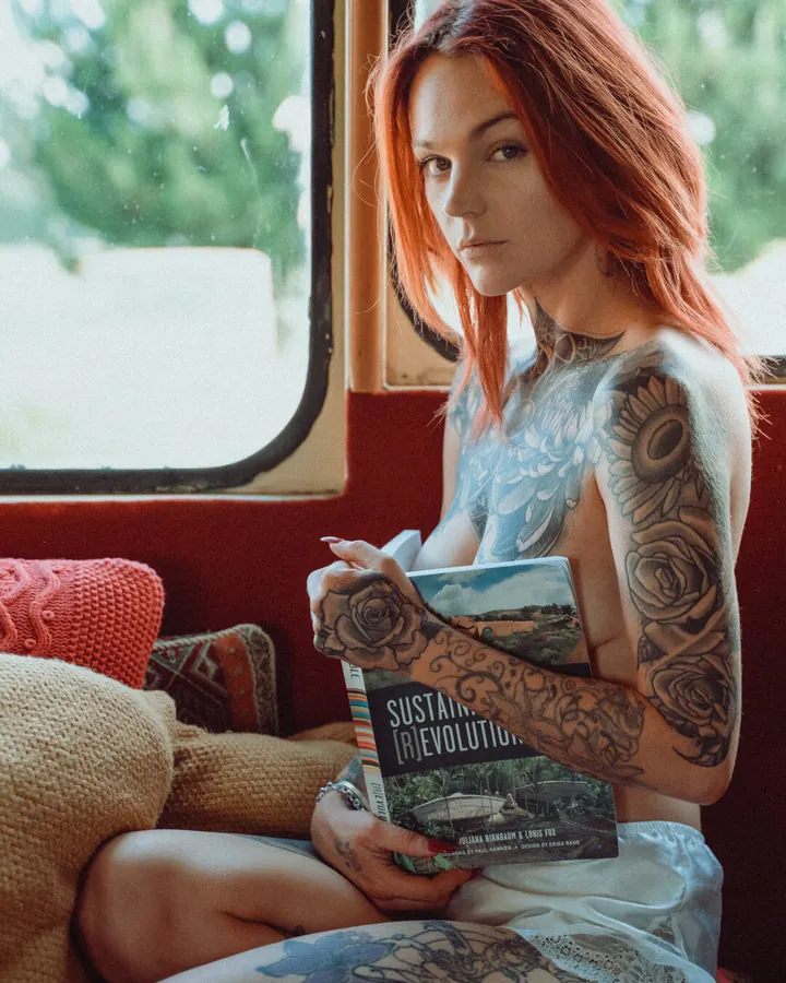 Femme aux cheveux roux, tatouages visibles sur le torse et les bras, assise dans un van, tenant un livre, regardant l'appareil.