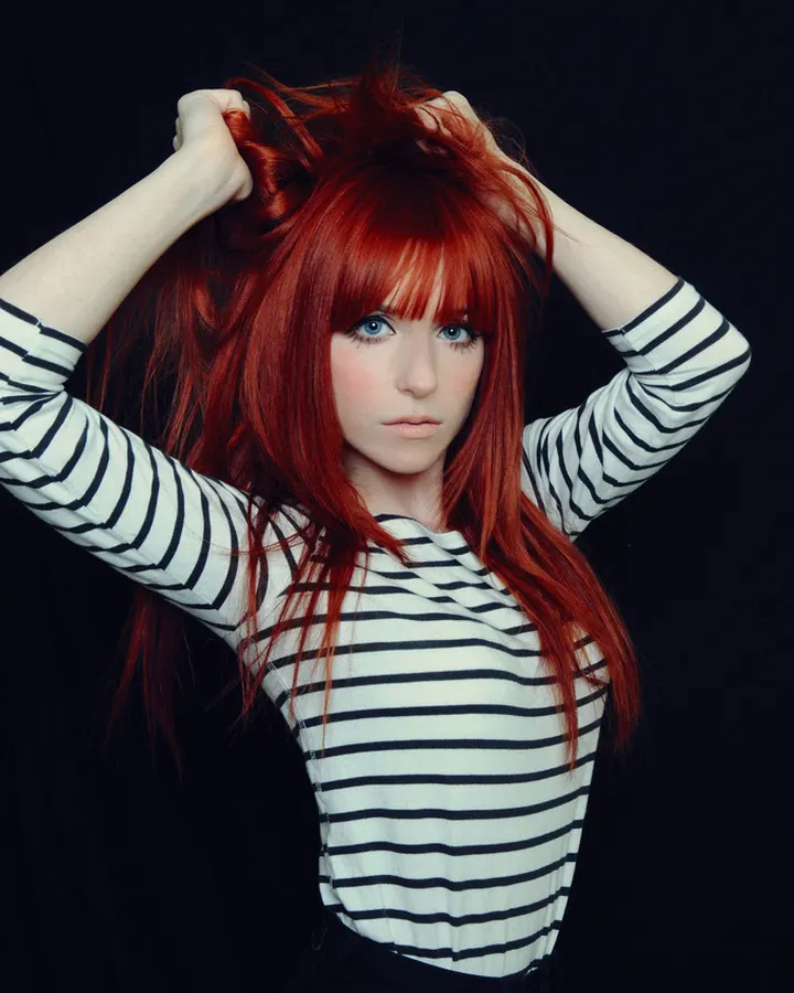 Femme aux cheveux roux, en haut rayé noir et blanc, tenant ses cheveux avec les mains, sur fond noir.