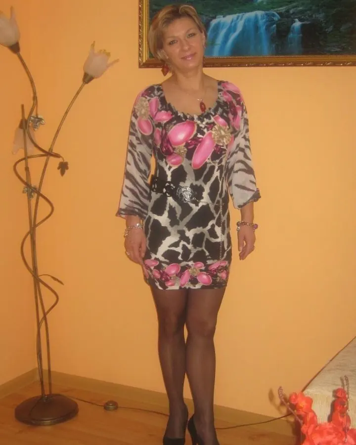 Femme en robe noire ajustée, posant dans une chambre avec lit et accessoires.