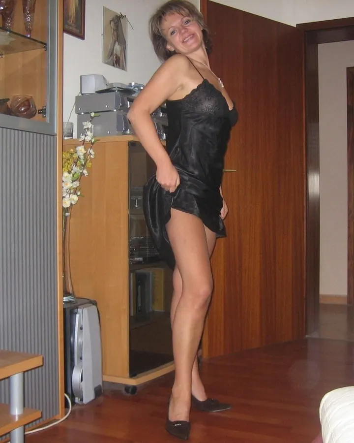 Femme en robe noire courte, posant dans un salon avec téléviseur et meubles.