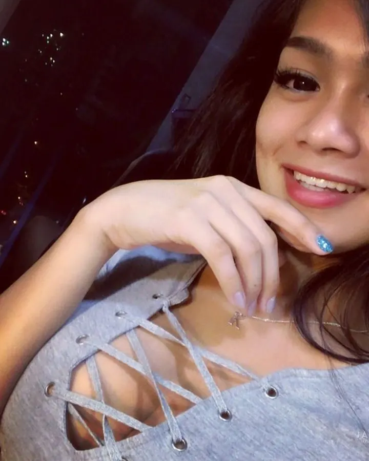 Jeune femme souriante en haut gris à lacets, posant près d'une fenêtre avec vue nocturne urbaine.