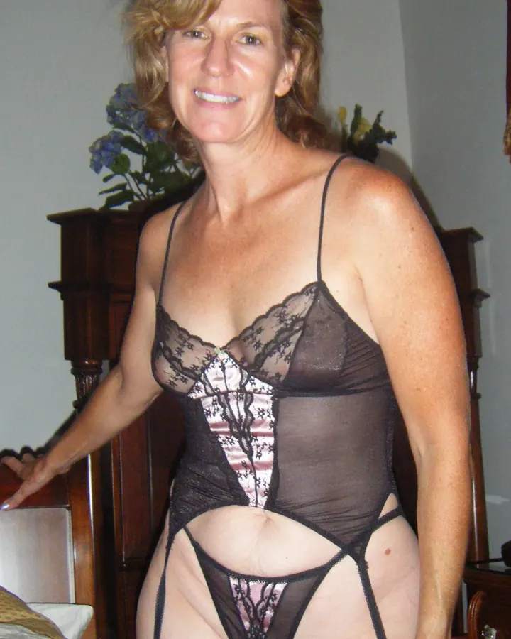 Femme blonde en lingerie noire transparente, debout près d'un lit dans une chambre intime.